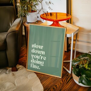 May include: A framed art print with a gold border and a teal background. The print features the words "slow down you're doing fine" in white text. The print is displayed on a wooden floor.