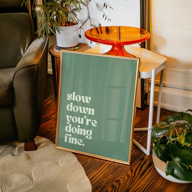 May include: A framed art print with a gold border and a teal background. The print features the words "slow down you're doing fine" in white text. The print is displayed on a wooden floor.