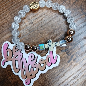 May include: A beaded bracelet featuring clear crackle glass beads, metallic rose gold spheres, iridescent square beads, and a dalmatian jasper cross charm. A gold-toned circular bead with a cross emblem is included. A sticker reads be the good.