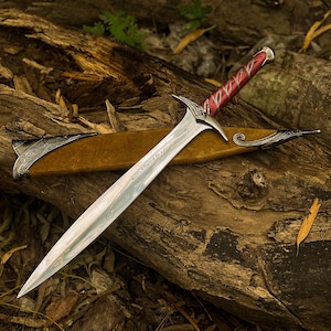 May include: A silver sword with a red handle and ornate detailing rests on a wooden log. The blade is sharp and the hilt is intricately designed. A tan sheath with silver accents is visible.