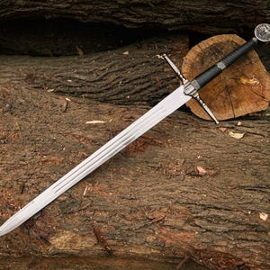 May include: A long, silver sword with a black handle and a decorative pommel rests on a pile of weathered logs. The blade has a central fuller and a crossguard. The sword is a medieval-style weapon.