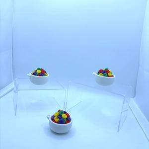 May include: Three small white ceramic bowls filled with colorful, round cereal pieces. The bowls are arranged on clear acrylic display stands against a light blue background. The cereal pieces are red, yellow, blue, and green.