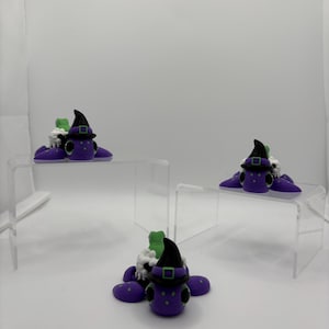 May include: Three miniature witch figurines. Each figurine is purple with a black hat and green accents. The figurines are displayed on clear acrylic risers against a white background. The witch figurines are a whimsical Halloween decoration.