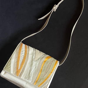 May include: A cream-colored shoulder bag with a flap closure. The bag features a decorative design with wavy patterns in shades of cream, gold, and light green. The strap is a matching cream color. The bag is set against a black background.