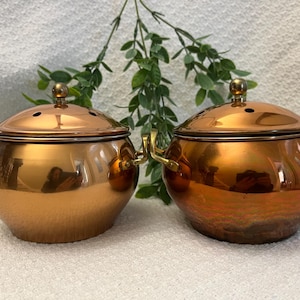 May include: Two copper-colored pots with lids. Each pot has a round body, a lid with a small knob, and two handles. The pots are reflective, showing the surrounding environment. The pots are sitting on a white surface.