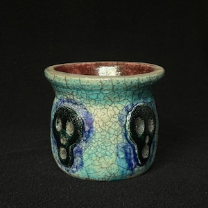 May include: A ceramic pot featuring a crackle glaze finish in shades of turquoise and blue. The exterior is decorated with two dark, glossy skull motifs. The interior of the pot is finished in a contrasting reddish-brown glaze with a wide rim.