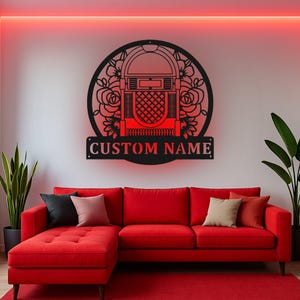 Op de afbeelding: Een rond wandbord van zwart metaal met een uitgesneden ontwerp van een jukebox omringd door bloemmotieven. Onder het cirkelvormige frame bevindt zich een rechthoekig plaatje met de tekst CUSTOM NAME in vetgedrukte, schreefloze hoofdletters.
