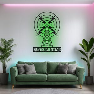 Peut inclure: Une décoration murale en métal noir représentant la silhouette d'une tour radio avec des éclairs et des ondes circulaires concentriques. Sous la tour, une barre horizontale affiche le texte CUSTOM NAME en lettres capitales.