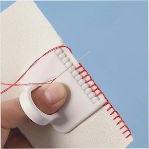 May include: A white plastic sewing tool with a finger ring, used for guiding the needle and thread. The tool is shown in use, with red thread and a needle stitching fabric. The background is a light blue.