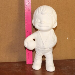May include: A white ceramic figurine of a smiling boy holding a football. The figurine is wearing a vest and shorts. A pink ruler is next to the figurine.