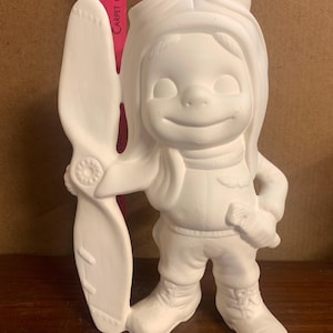 Puede incluir: Figurilla de cerámica blanca de un niño con un gorro y gafas de piloto, sosteniendo una hélice. La figurita está lista para ser pintada.