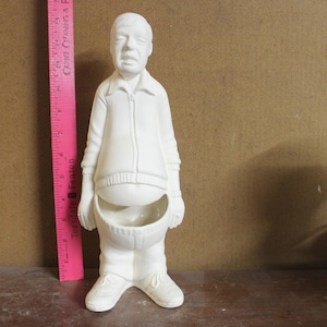May include: A white ceramic figurine of a man wearing a zip-up jacket, sweater, and pants. The figurine has a large belly with a bowl-shaped opening. The man is wearing sneakers. A pink ruler is next to the figurine.
