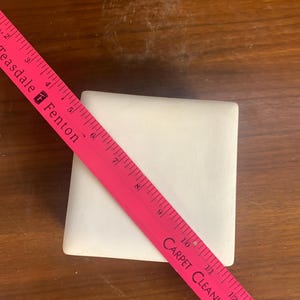 May include: A white square ceramic tile measuring 10 inches by 10 inches. The tile is sitting on a wooden surface. A pink ruler is placed diagonally across the tile.