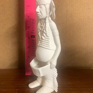 Puede incluir: Una figura de cerámica blanca de un hombre nativo americano, con un tocado y sosteniendo una bolsa. La figura mide aproximadamente 20 cm de altura, según la regla rosa del fondo. La figura está sobre una superficie de madera.