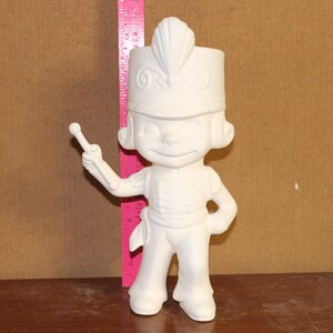 May include: A white ceramic toy soldier figurine, wearing a hat and uniform, holding a baton. The figurine is posed with one hand on its hip. A pink ruler is positioned behind the figurine. The figurine is unpainted.
