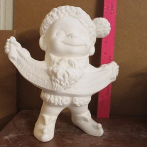 May include: A white ceramic figurine of a character wearing a hat and boots, holding a banner that reads Merry Christmas. A pink ruler is positioned behind the figurine, showing a height of approximately 8 inches for the ceramic object.