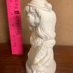 Puede incluir: Figurilla de cerámica blanca de una niña con el pelo largo que lleva un sombrero y un vestido. La figurita mide aproximadamente 15 cm de alto.
