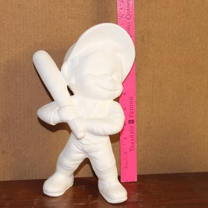 Puede incluir: Figura de cerámica blanca de un jugador de béisbol en posición de bateo, sosteniendo un bate. La figura lleva gorra y uniforme de béisbol. Una regla rosa está colocada junto a la figura. La figura no está pintada.