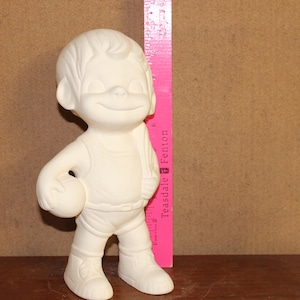 May include: A bisque ceramic figurine of a smiling boy holding a ball, with a pink ruler next to it. The figurine is wearing a tank top, shorts, and sneakers. The ruler is approximately 12 inches tall.