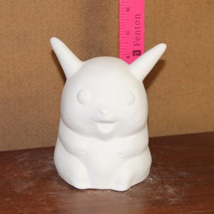 May include: A white ceramic figurine of a cartoon character with large ears and a smiling face. The figurine is unpainted and appears to be a blank canvas for customization. A pink ruler is placed behind the figurine.