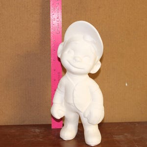 May include: A white ceramic figurine of a smiling boy wearing a baseball cap and a vest. The figurine is standing next to a pink ruler. The figurine is approximately 7 inches tall.