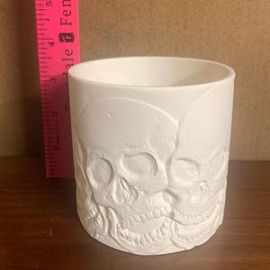 Könnte beinhalten: Weißer Keramik-Pflanztopf mit Totenkopf-Design. Der Topf ist zylindrisch und hat eine glatte, matte Oberfläche. Die Totenköpfe sind kreisförmig um den Topf angeordnet.