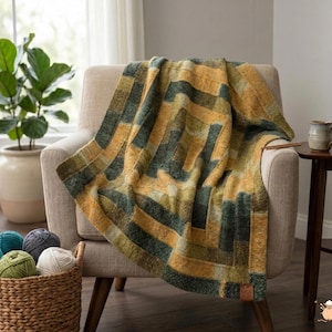 May include: A cozy, geometric-patterned blanket in shades of gold, green, and olive green is draped over a beige armchair. The blanket features a textured design and is made of soft material. A basket of yarn balls sits nearby.