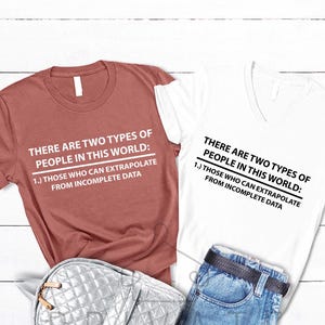 May include: Two t-shirts, one brick red and one white, with the text "THERE ARE TWO TYPES OF PEOPLE IN THIS WORLD: 1.) THOSE WHO CAN EXTRAPOLATE FROM INCOMPLETE DATA." A silver quilted bag and blue jeans are also in the image.
