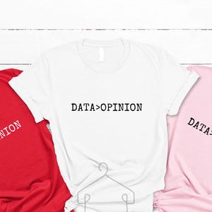 May include: Three t-shirts in red, white, and pink, each with the text "DATA>OPINION" printed in black. The shirts are displayed on a white wooden surface.