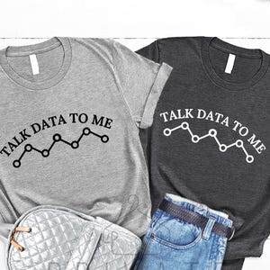 May include: Two t-shirts, one gray and one dark gray, with the text "TALK DATA TO ME" and a line graph design. The shirts are paired with blue jeans and a silver quilted bag on a white wooden surface.
