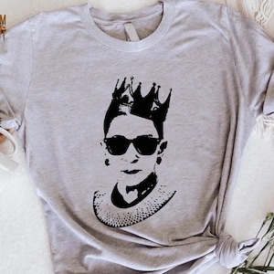 May include: A gray t-shirt with a black silhouette of Ruth Bader Ginsburg wearing a crown and glasses.
