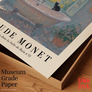 May include: A framed art print of Claude Monet's painting, "Dans la Salle de Bain n&deg;11." The artwork shows a bathtub, a black cat, flowers, and a window. The frame is light-coloured wood. The print is on museum-grade paper.