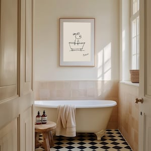 May include: A framed art print featuring a minimalist line drawing of a dog in a bathtub. The artwork is in black on a cream background, with the artist's signature. The bathroom has a clawfoot tub, a wooden stool, and a black and white checkered floor.