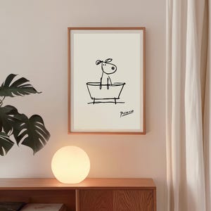 May include: A framed art print featuring a minimalist black line drawing of a dog in a bathtub. The artwork is signed "Picasso" in the lower right corner. The print is displayed on a light-colored wall above a wooden cabinet with a round, glowing lamp.