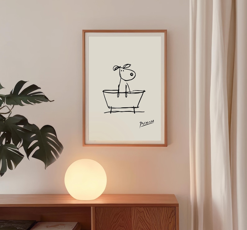 May include: A framed art print featuring a minimalist black line drawing of a dog in a bathtub. The artwork is signed "Picasso" in the lower right corner. The print is displayed on a light-colored wall above a wooden cabinet with a round, glowing lamp.