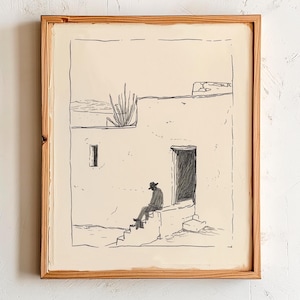 May include: A framed black and white ink drawing depicts a person seated on steps outside a building. The artwork features a desert landscape with a cactus and a door. The frame is made of light-colored wood.