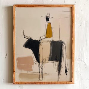 Könnte beinhalten: Abstrakter Kunstdruck in einem Holzrahmen. Das Kunstwerk zeigt eine Figur in einem gelben Hemd und schwarzem Hut, die einen schwarz-weißen Bullen reitet. Der Hintergrund weist neutrale Beige- und Brauntöne auf.