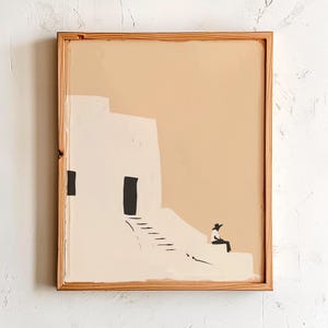 May include: Abstract art print in a wooden frame. The artwork depicts a minimalist scene of a white building with a black door and a person sitting on a ledge. The background is a warm beige color.
