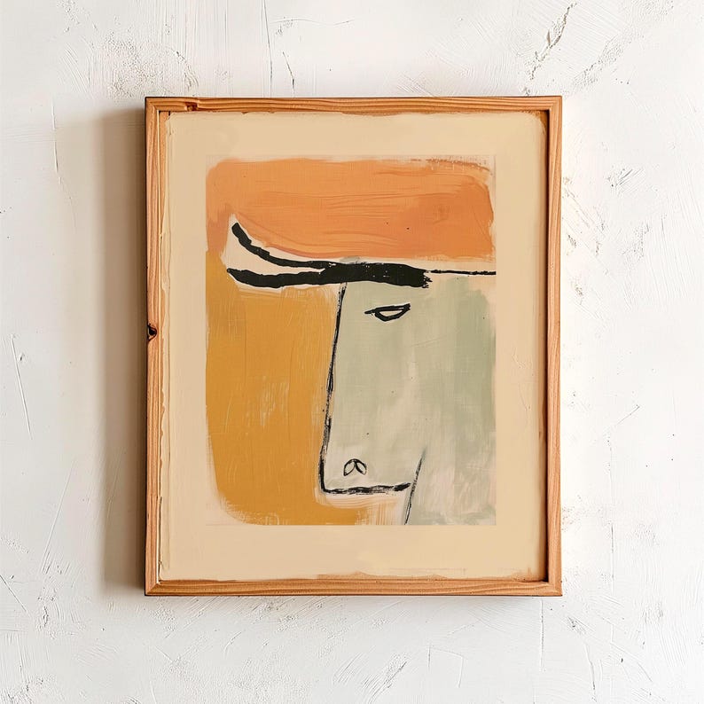 Op de afbeelding: Abstracte kunstprint met een gestileerd gezicht in groen, oranje en zwart. Het kunstwerk is ingelijst in een natuurlijke houten lijst. Het schilderij heeft een minimalistische stijl en een moderne esthetiek.