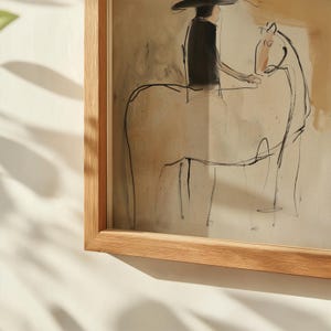 May include: Abstract artwork in a light wood frame. The artwork depicts a person on a horse, rendered in black lines against a beige background. The person is wearing a hat and dark clothing. The horse is drawn with simple lines.