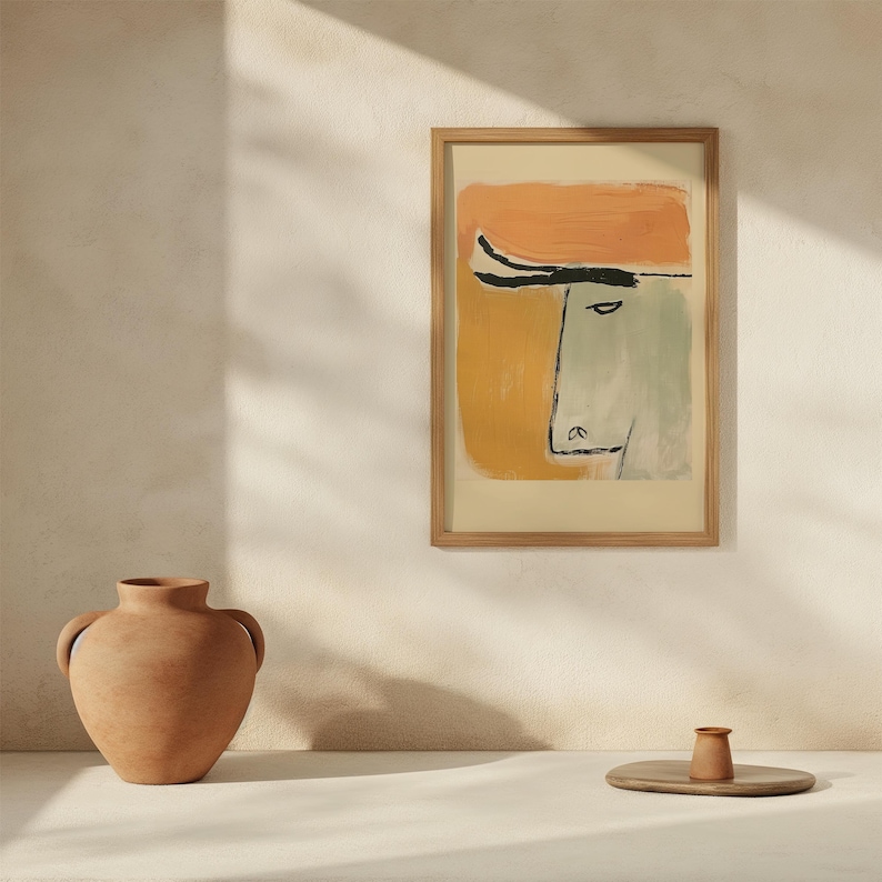 Op de afbeelding: Abstracte kunstprint in een houten lijst. Het kunstwerk toont een gezicht met een oranje hoed en een gele en groene achtergrond. Een terracotta vaas en een kleine houten dienblad met een terracotta kopje staan op de voorgrond.