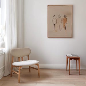 Puede incluir: Un interior minimalista con una silla de madera clara con asiento y respaldo crema, una peque&ntilde;a mesa auxiliar de madera y una obra de arte enmarcada que representa tres figuras con sombreros. La habitaci&oacute;n tiene pisos de madera y una paleta de colores neutros.