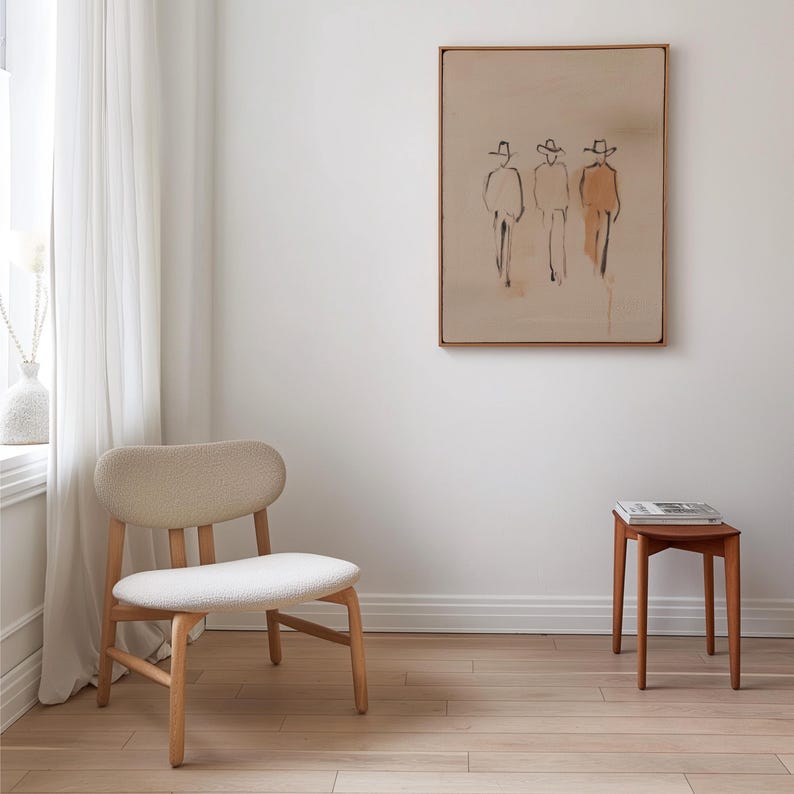 Puede incluir: Un interior minimalista con una silla de madera clara con asiento y respaldo crema, una peque&ntilde;a mesa auxiliar de madera y una obra de arte enmarcada que representa tres figuras con sombreros. La habitaci&oacute;n tiene pisos de madera y una paleta de colores neutros.