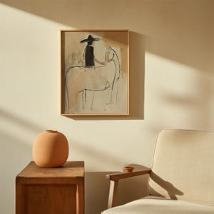 May include: A framed artwork depicts a person on a horse, rendered in a minimalist style with black and beige tones. The artwork is in a light wooden frame, hanging on a beige wall. A terracotta vase sits on a wooden side table, with a small bowl on the arm of a chair.