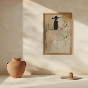 May include: A framed artwork depicts a person in a hat riding a horse, rendered in a minimalist style with black and beige tones. The artwork is in a light wood frame. A terracotta vase and a small wooden tray with a small terracotta cup are in the foreground.