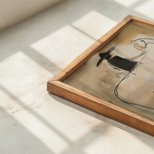 May include: A framed artwork featuring a sketch of a person wearing a hat and dress. The artwork is in a wooden frame. The sketch is in black and the background is beige and white. The frame is made of light brown wood.