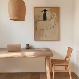 May include: A neutral-toned room featuring a wooden desk with a laptop, a small vase, and a framed artwork depicting a person on a horse. A woven light fixture hangs above the desk, and a woven chair sits nearby. The artwork is the focal point.