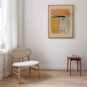 Op de afbeelding: Een abstracte kunstprint van een stierenkop in een houten lijst hangt aan een witte muur. Het kunstwerk heeft oranje, gele en groene kleuren. Een lichtgekleurde stoel en een kleine houten bijzettafel staan in de kamer.