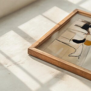 May include: A framed abstract art piece with a light wood frame. The artwork features black, white, and yellow brushstrokes on a beige background. The frame is rectangular and made of natural wood.