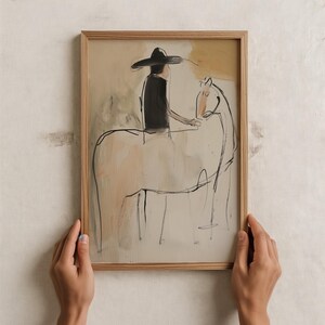 May include: Abstract art print in a natural wood frame. The artwork depicts a person in a hat riding a horse, rendered in black and neutral tones. The hands holding the frame are visible.